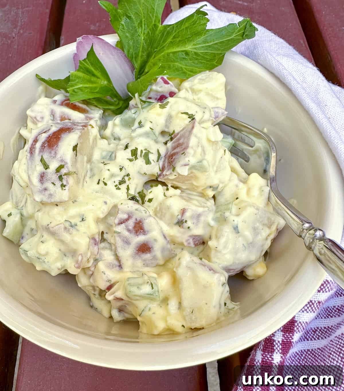 Creamy Red Potato Salad with Herbs and Dijon 2 Red potato salad in a bowl with a sprig of celery, a fork in the bowl and a napkin on the side.