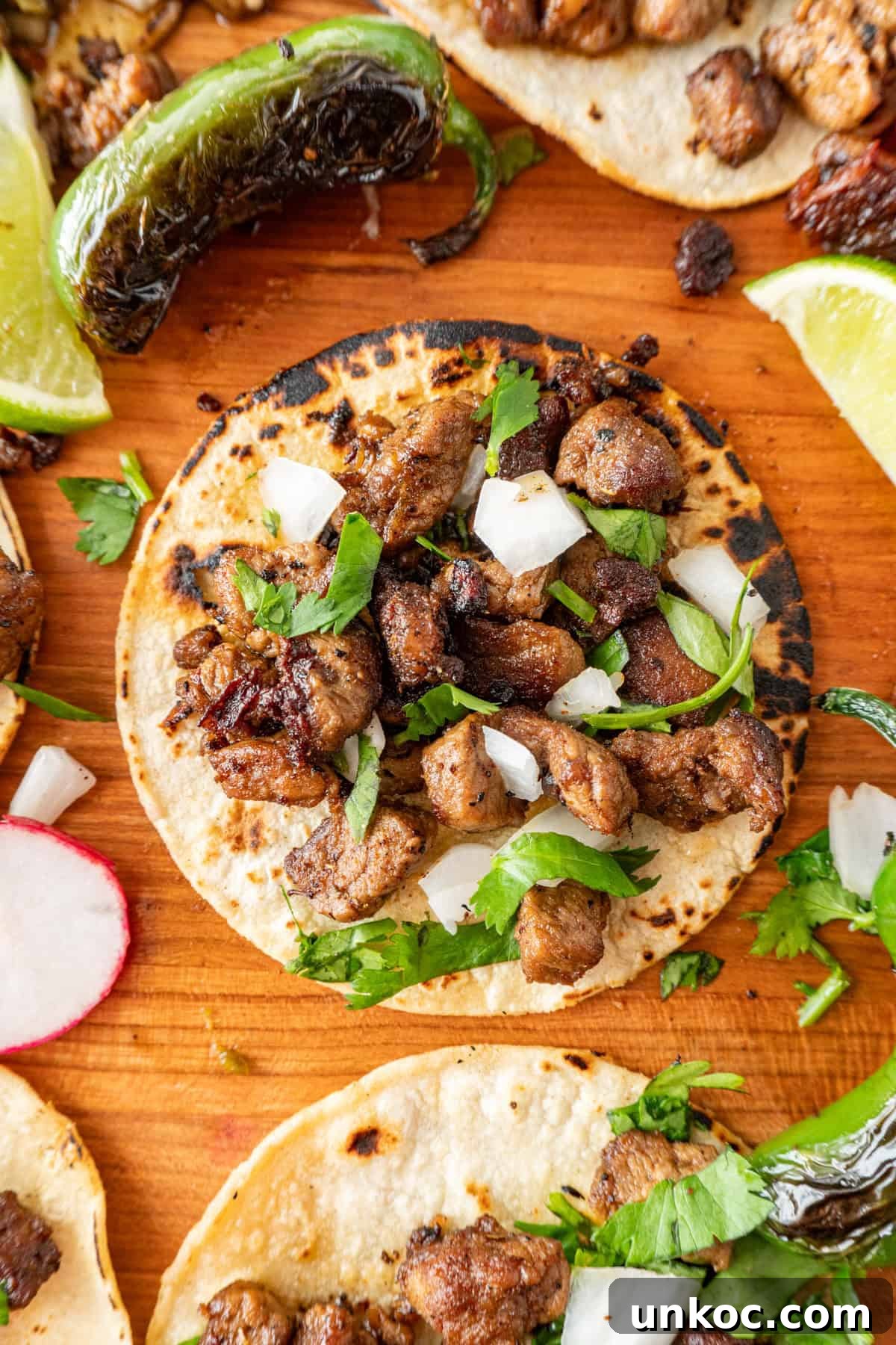 Taco sprinkled with onion and cilantro on a cutting board surrounded by blackened jalapeno and lime wedges.