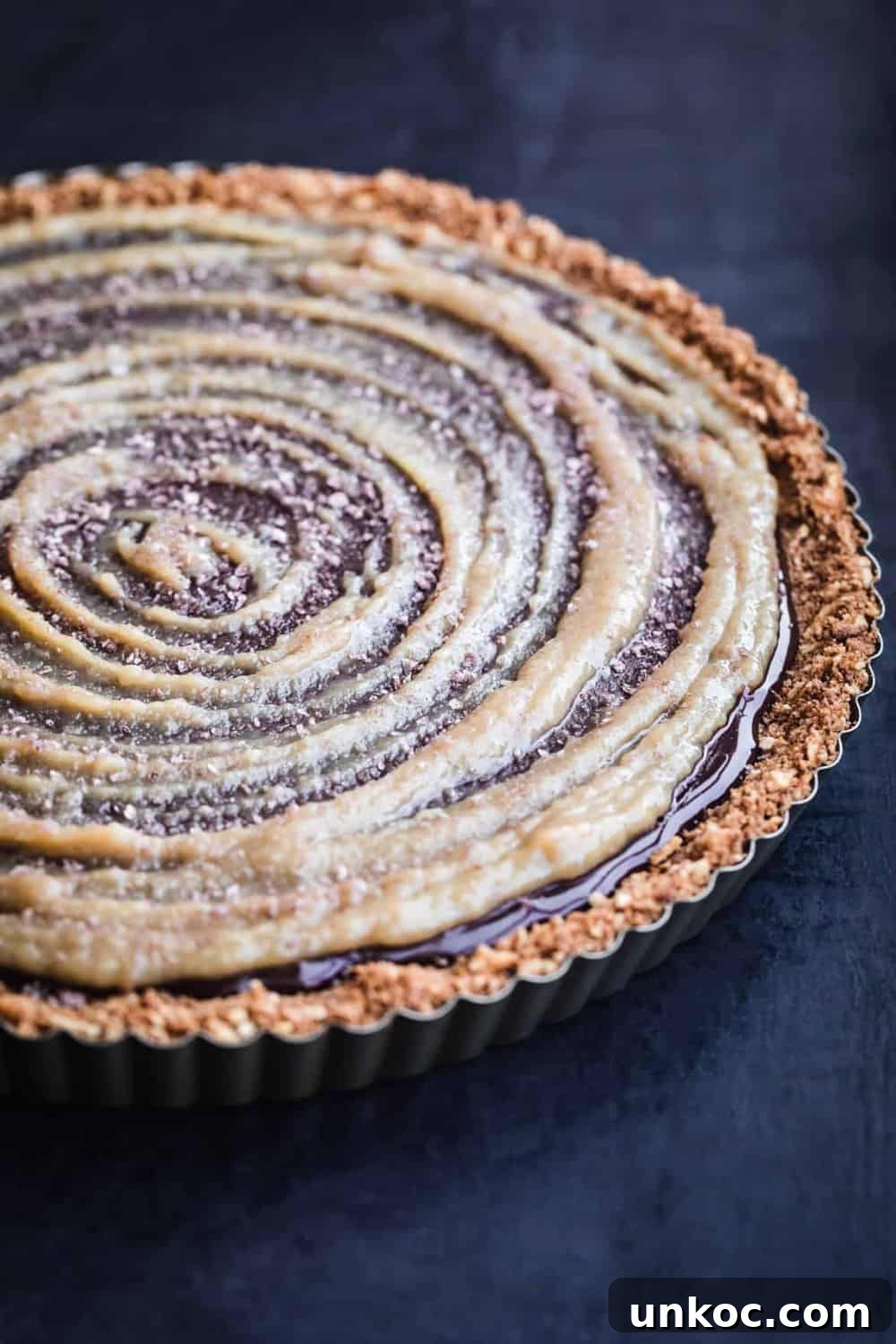 Chocolate Tahini Tart Recipe with Silky Ganache 2 Chocolate tahini tart with a glossy dark chocolate ganache filling, tahini swirl on top and a pretzel crust in a tart pan on a dark background.
