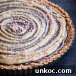 Chocolate Tahini Tart Recipe with Silky Ganache 14 Chocolate tahini tart with a glossy dark chocolate ganache filling, tahini swirl on top and a pretzel crust in a tart pan on a dark background.