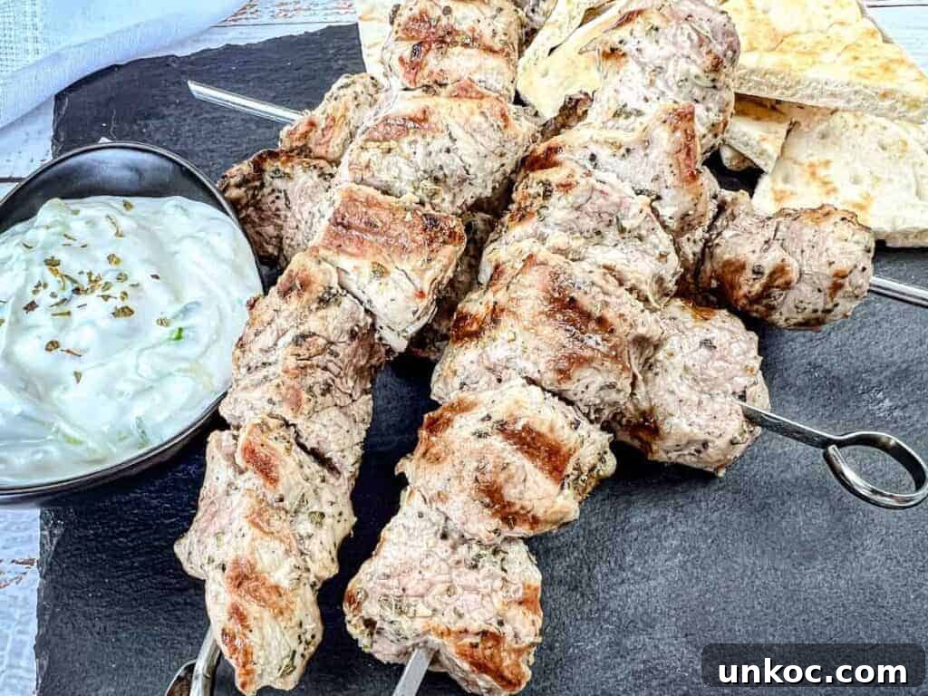 Three pieces of Pork Souvlaki with a bowl of white sauce and pieces of flatbread on a black slate serving board.
