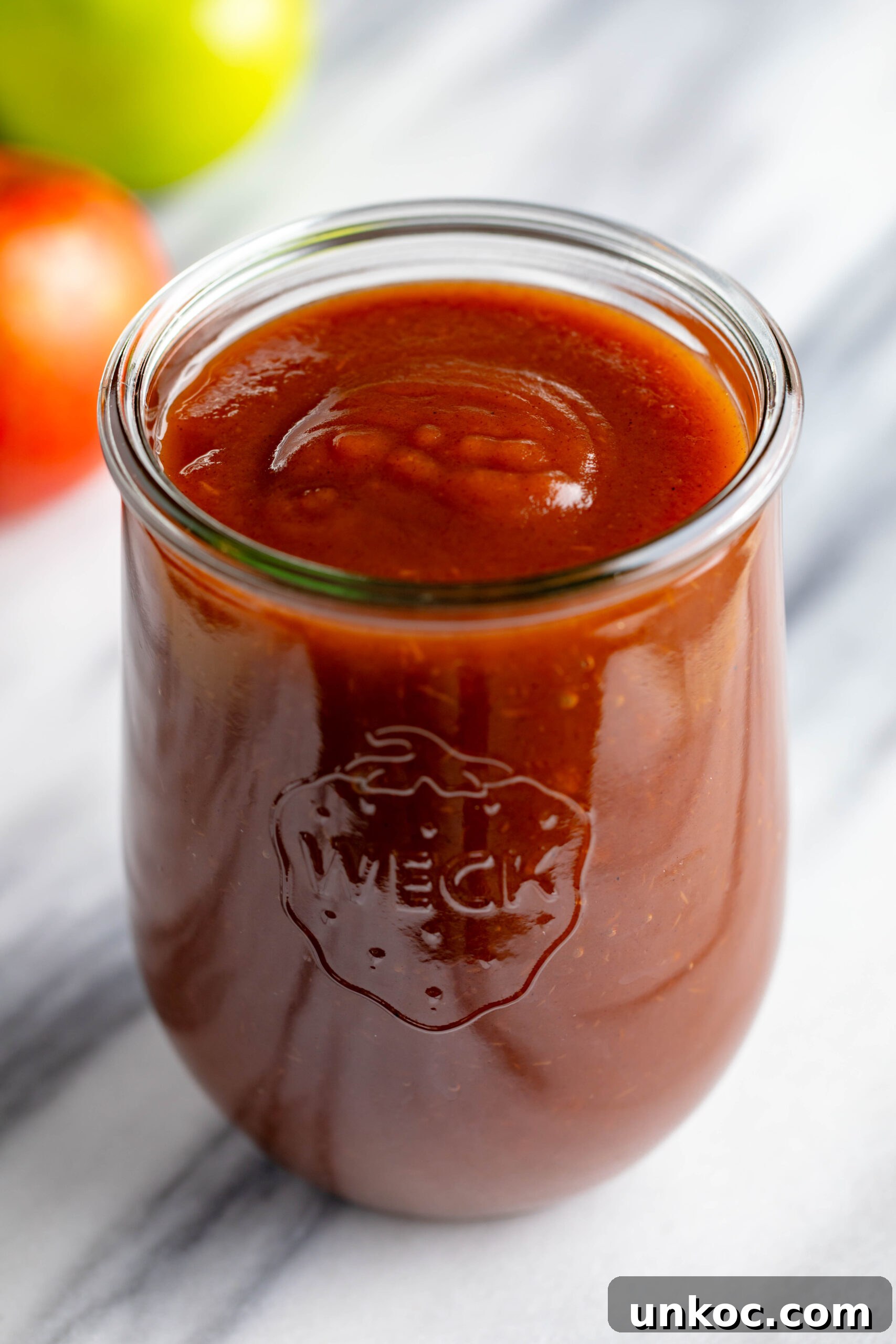 Instant Pot Apple Butter Recipe No Peeling Required 12 weck jar filled with apple butter on a white marble slab with a red and green apple in the background